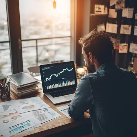 فردی در حال بررسی نمودارهای اقتصادی و تجاری روی لپ‌تاپ در یک محیط کاری، با مستندات و گزارش‌های چاپ‌شده روی میز.